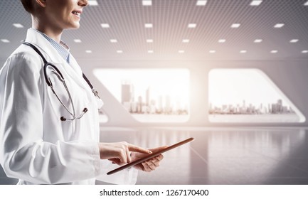 Side View Of Young Female Medical Industry Employee Holding Tablet In Hands While Standing Inside Bright Hospital Building. Medical Industry Concept