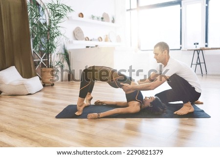 Side view of young female standing on leg toes and supporting hands while lifting waist up in backbend exercise with help of instructor at home