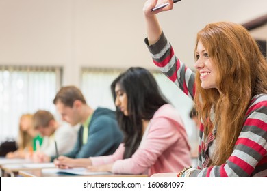 Side View Of A Young Female Student Raising Hand By Others In The Classroom