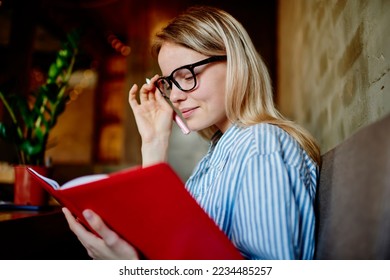 Side view of young female student adjusting eyeglasses with hand while sitting at table with smartphone coffee cup and reading notes in notepad in blurred cafe - Powered by Shutterstock