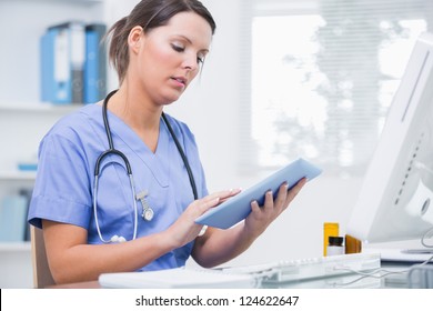 Side view of young female surgeon using digital tablet in front of computer at desk in clinic
