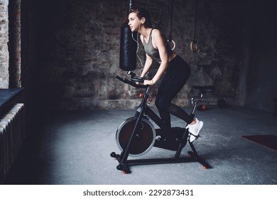 Side view of young fit female with device on arm and earphones doing cardio practice on stationary exercise bicycle in gym