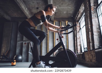 Side view of young fit female with device on arm and earphones doing cardio practice on stationary exercise bicycle in gym - Shutterstock ID 2292873439