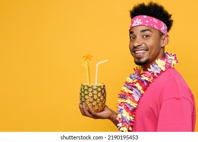 SIde View Young Fun Man 20s He Wearing Pink T-shirt Hawaiian Lei Near Hotel Pool Drink Straw Pineapple Cocktail Juice Look Camea Isolated On Plain Yellow Background. Summer Vacation Sea Rest Concept