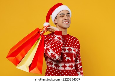 Side view young fun man wear red knitted christmas sweater Santa hat hold open look at shopping paper package bags isolated on plain yellow background. Black Friday New Year 2023 sale buy day concept