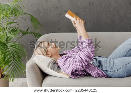 Side view young fun smart woman wearing casual clothes reading book novel lying on grey sofa couch stay at home hotel flat rest relax spend free spare time in living room indoor. People lounge concept