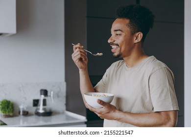 Side view young fun smiling african american man in casual clothes eat breakfast muesli cereals with milk fruit in bowl prepare cook food in light kitchen at home alone indoor. Healthy diet concept. - Powered by Shutterstock