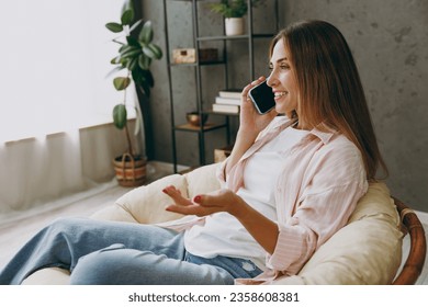 Side view young fun woman wear casual clothes talk speak on mobile cell phone sits in armchair stay at home hotel flat rest relax spend free spare time in living room indoor. Lifestyle lounge concept