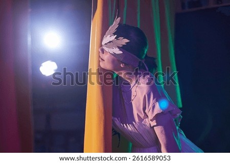 Side view of young girl dressed in handmade paper costume with feather headband peeking from behind yellow curtains during fairytale play at childrens theater waiting to step on stage, copy space
