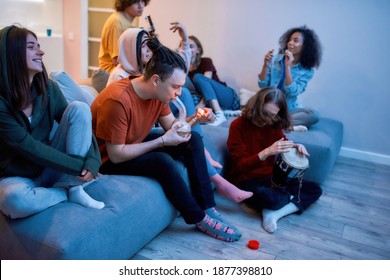 Side View Of A Young Guy Lighting Marijuana In The Glass Bong, Relaxing With Friends On The Sofa At Home, Young People Smoking Weed Together. Man Playing Bongo. Cannabi, Ganja, Marijuana Home Party