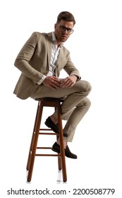 Side View Of Young Guy In Suit With Open Collar Shirt Sitting On Wooden Chair And Confidently Posing On White Background In Studio