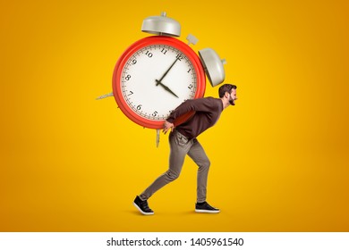 Side view of young handsome man carrying huge alarm clock on his back on yellow gradient background. Meet deadlines. Live in hectic world. Adjust to overbusy schedule.