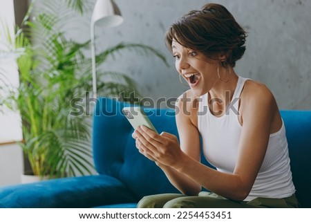 Side view young happy shocked woman wear white tank shirt hold use mobile cell phone sit on blue sofa stay at home flat rest relax spend free spare time in living room indoors People lounge concept