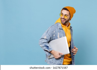 Side View Of Young Hipster Guy In Fashion Jeans Denim Clothes Posing Isolated On Pastel Blue Background In Studio. People Lifestyle Concept. Mock Up Copy Space. Hold Laptop Pc Computer, Looking Back