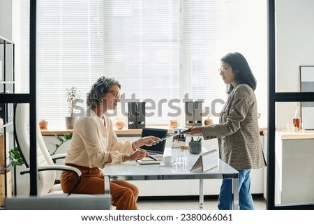 Side view of young Hispanic female white collar worker passing document to boss or ceo of business company to check main points of contract