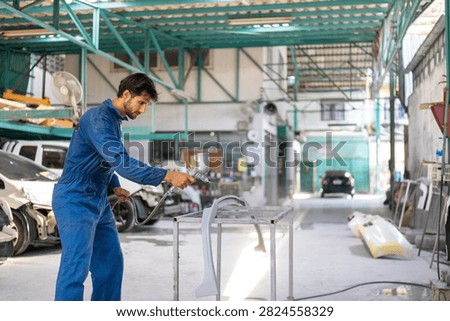 Side view of a young Hispanic mechanic in blue coveralls spray painting a car bumper on a metal rack.