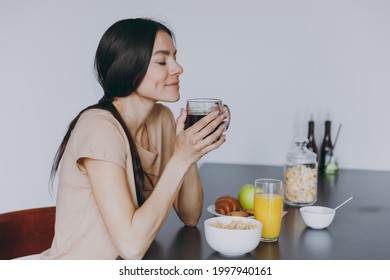 Side View Young Housewife Woman 20s Wear Casual Clothes Beige Shirt Eat Breakfast Hold Cup Of Tea Drink Coffee Sniff Scent Cooking Food In Light Kitchen At Home Alone. Healthy Diet Lifestyle Concept