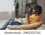 Side view young Indian man wear orange casual clothes reading novel book drink coffee sits in armchair stay at home hotel flat rest relax spend free spare time in living room indoor. Lounge concept