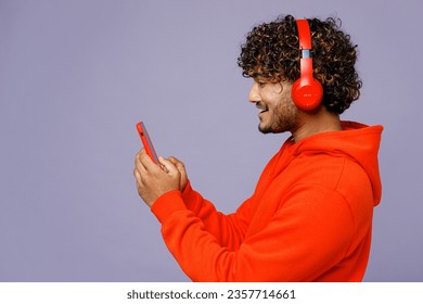 Side view young Indian man wear red orange hoody casual clothes listen to music in headphones use mobile cell phone isolated on plain pastel purple color background studio portrait. Lifestyle concept - Powered by Shutterstock