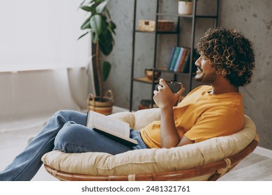 Side view young Indian man wear orange casual clothes reading novel book drink coffee sits in armchair stay at home hotel flat rest relax spend free spare time in living room indoor. Lounge concept - Powered by Shutterstock