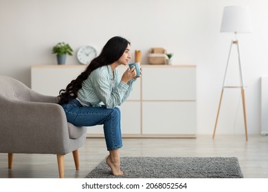 Side view of young Indian woman sitting in armchair, sipping hot aromatic coffee, enjoying lazy relaxing day at home, copy space. Lovely Eastern lady drinking warm beverage indoors - Powered by Shutterstock