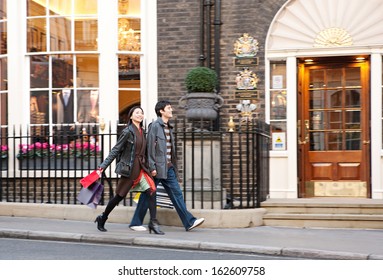 Side View Of A Young Japanese Tourist Couple On Holiday Walking Down An Exclusive And Luxurious Shopping Street In London City With Classic Stores And Holding Carrier Paper Bags, Outdoors.