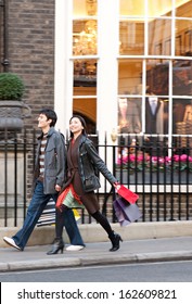 Side View Of A Young Japanese Tourist Couple On Vacation Walking Down An Exclusive And Luxurious Shopping Street In London City With Classic Stores And Holding Carrier Paper Bags, Outdoors.