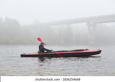 Side View Of Young Kayaker In River, Padding, Man Enjoying Water Sport In Foggy Morning With Bridge On Background, Male Rowing Boat In Cold Autumn Day.