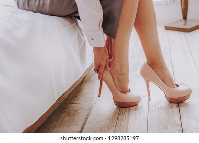 Side View Of Young Lady Putting On Stilettos While Sitting On Bed In Apartment. Focus On Her Legs In Shoes