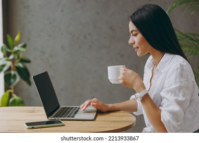 Side view young latin IT woman in white shirt hold use work or study on laptop pc computer drink tea sit alone at table in coffee shop cafe restaurant indoors Freelance mobile office business concept - Powered by Shutterstock