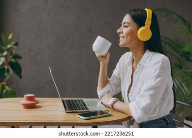 Side view young latin IT woman in white shirt headphones listen music hold use work study laptop pc computer drink coffee sit at table in coffee shop cafe restaurant indoor Freelance business concept - Powered by Shutterstock