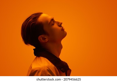 Side view of young male with closed eyes tilting back head while standing under bright neon illumination against orange background