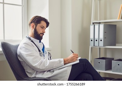 Side View Of Young Male Doctor Writing Case Report In Comfortable Medical Office.