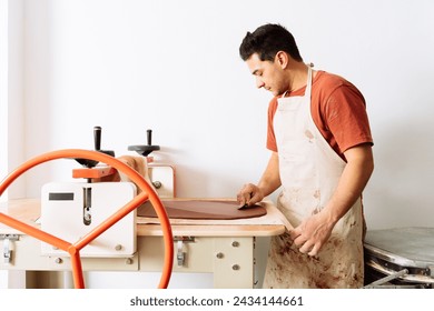 Side view of young male potter in dirty apron standing at slab roller while working with clay in professional pottery workshop - Powered by Shutterstock