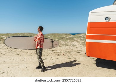 Side view of young male surfer in casual clothes and sunglasses walking on sandy beach near retro camper van with surfboard in hand - Powered by Shutterstock