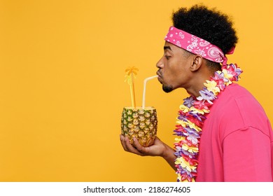 SIde View Young Man 20s He Wearing Pink T-shirt Hawaiian Lei Near Hotel Pool Drink Straw Pineapple Cocktail Juice Isolated On Plain Yellow Background Studio Portrait. Summer Vacation Sea Rest Concept
