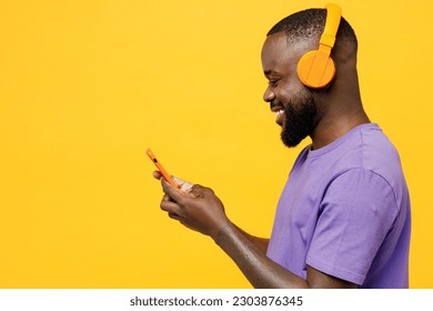 Side view young man of African American ethnicity wear casual clothes purple t-shirt headphones listen to music hold use mobile cell phone isolated on plain yellow background studio. Lifestyle concept - Shutterstock ID 2303876345
