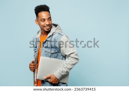 Side view young IT man of African American ethnicity wear denim jacket orange t-shirt hold closed laptop pc computer look aside isolated on plain pastel light blue cyan background. Lifestyle concept