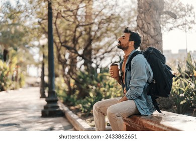 Side view of a young man with backpack sitting on a city street downtown and enjoying his takeaway coffee.