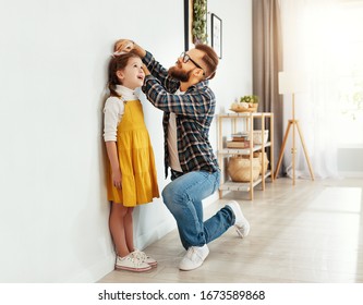 Side view of young man in casual clothes measuring height of little daughter next to white wall in light living room