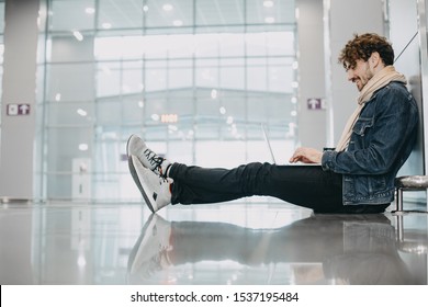 Side view of young man with curly hair sitting on floor and waiting for his flight in airport. Work remote. Holding laptop on nap and typing. Smile. Alone in hall. Modern view - Powered by Shutterstock