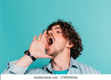 Side Up View Of Young Man With Curly Hair And Beard Screaming Out Loud. Holding Hand Close To Mouth. Shout In The Air. Isolated Over Blue Background