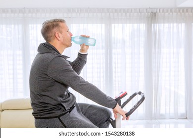 Side View Of Young Man Drinking A Bottle Of Water While Training On A Spin Bike In The Living Room. Shot At Home