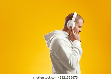 Side view of young man enjoying music through wireless headphones with closed eyes and touching headset with fingers against yellow background - Powered by Shutterstock