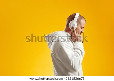 Side view of young man enjoying music through wireless headphones with closed eyes and touching headset with fingers against yellow background