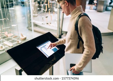 Side view of young man in glasses with black backpack holding laptop and smartphone while using information digital stand in hall. Male using informative electronic kiosk