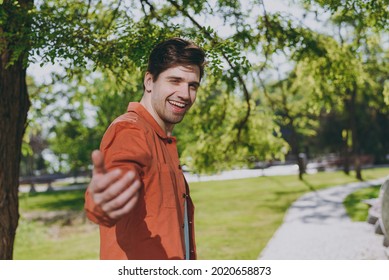 Side View Young Man In Orange Jacket Walking Stretch Hand To Camera Say Come With Me Call Rest Relax In Spring Green City Park Go Down Alley Sunshine Lawn Outdoors On Nature. Urban Leisure Concept.