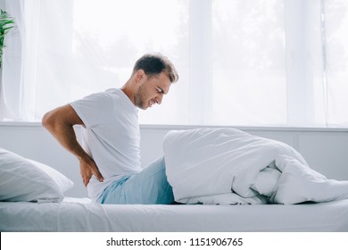 Side View Of Young Man In Pajamas Sitting On Bed And Suffering From Back Pain