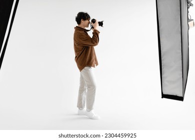 Side view of young man photographer taking photo using professional dslr camera and lighting equipment, standing on white background, copy space, full length - Powered by Shutterstock