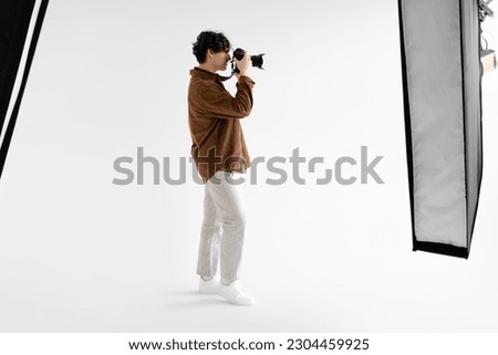 Side view of young man photographer taking photo using professional dslr camera and lighting equipment, standing on white background, copy space, full length
